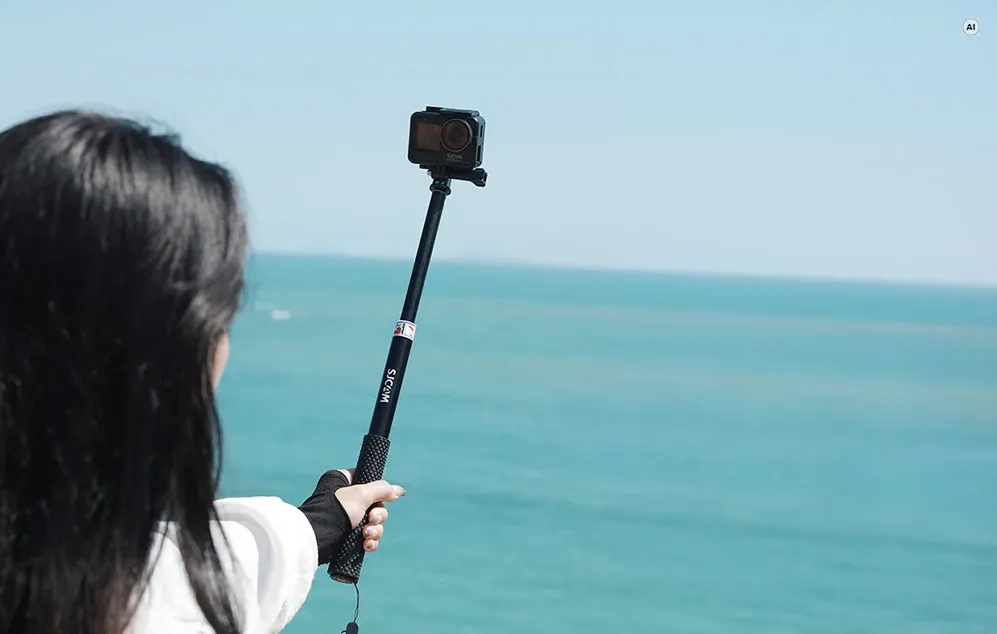 O femeie cu părul lung și negru ține un selfie stick cu o cameră de acțiune, având marea ca fundal