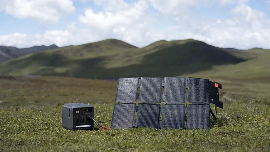 Un panou solar portabil așezat pe iarbă este conectat printr-un cablu la stația de alimentare DJI Power 1000 Mini, pe fundalul unor dealuri verzi