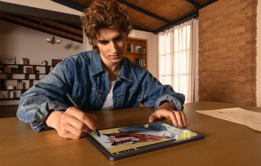 O persoană îmbrăcată într-o jachetă din denim desenează o ilustrație colorată cu un stilou pe o tabletă așezată pe o masă