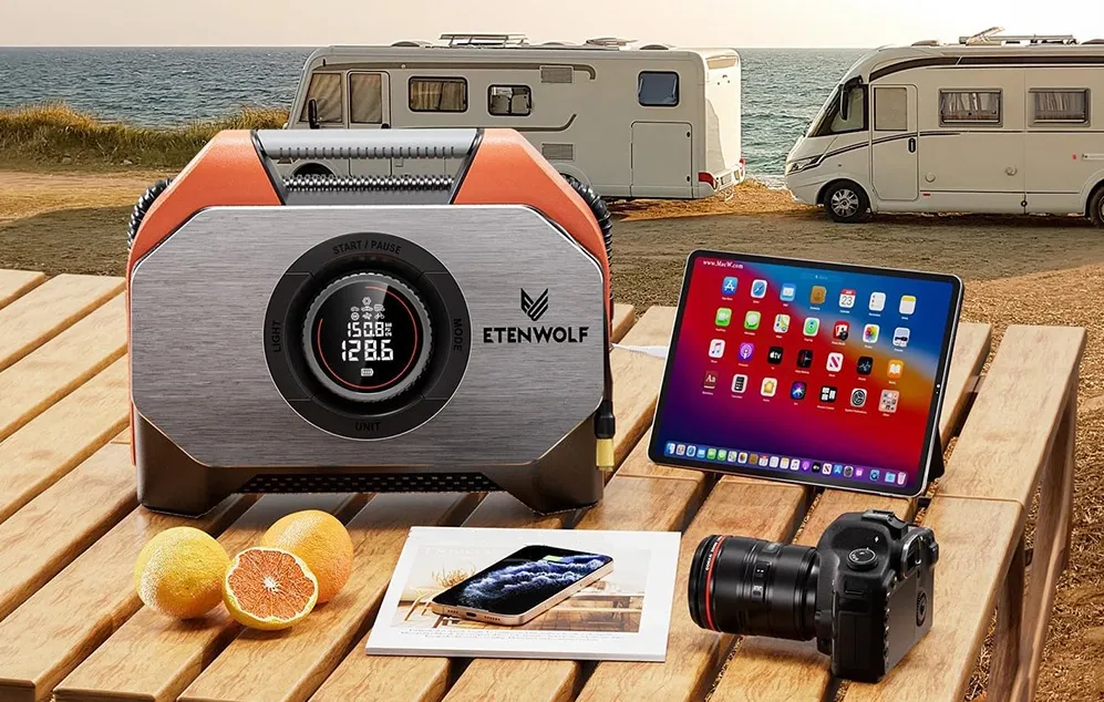 Un compresor Etenwolf pe o masă de lemn lângă o tabletă, un smartphone, un aparat foto și un ziar, cu două rulote pe malul unui lac în fundal