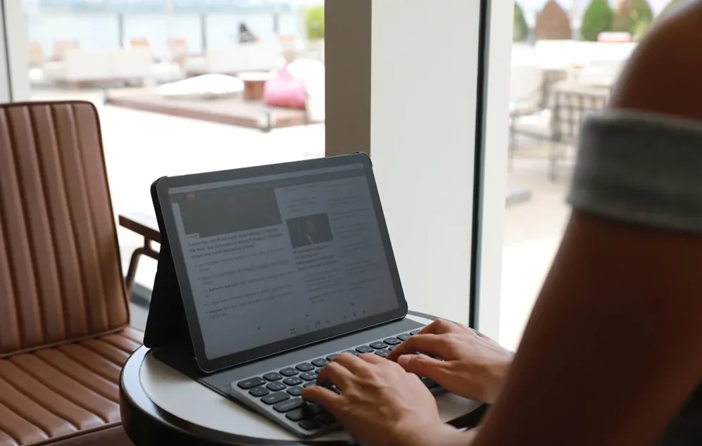 O persoană tastează pe o tastatură conectată la o tabletă așezată pe o măsuță lângă o fereastră mare