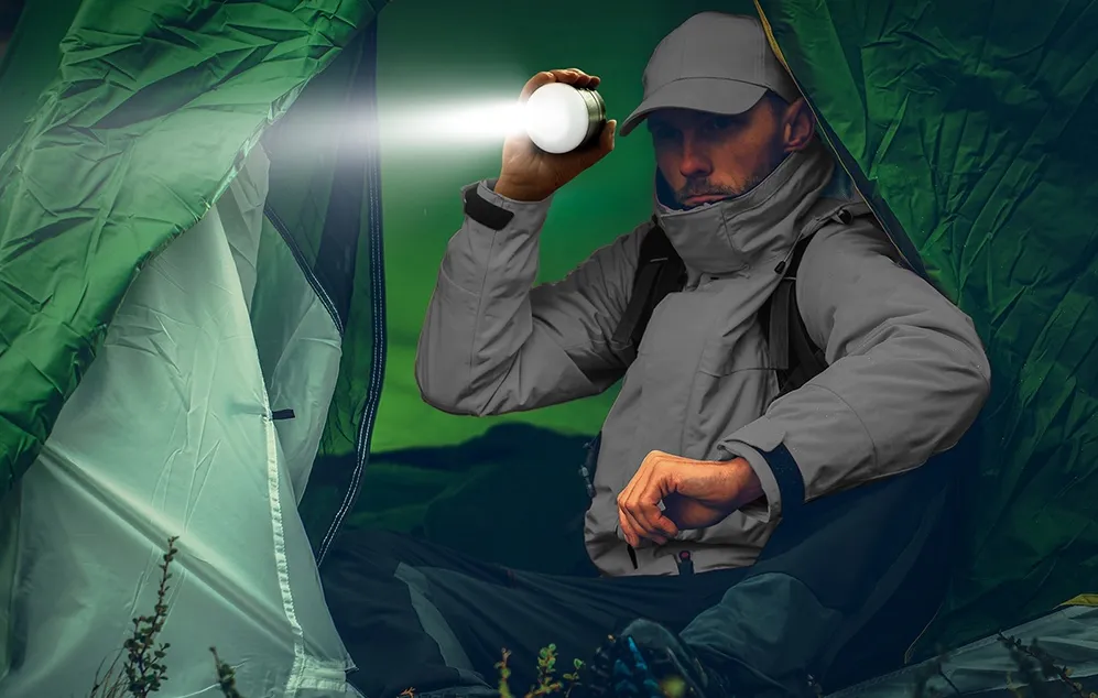 Un bărbat îmbrăcat într-o jachetă gri stă într-un cort verde, îndreptând în fața sa lumina unei lămpi rotunde pe care o ține în mână.