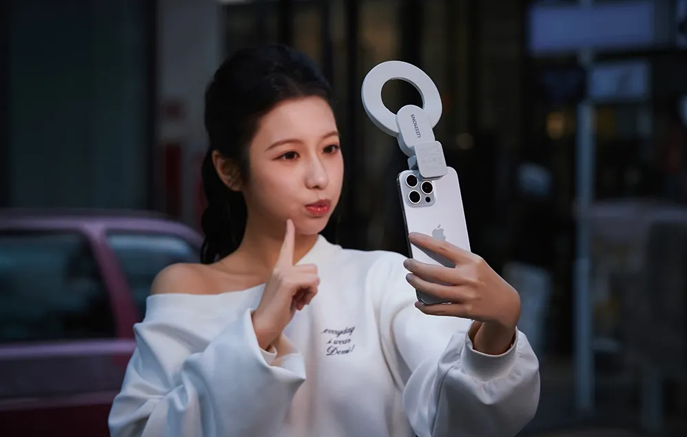 O femeie care își face un selfie cu smartphone-ul, având atașată o lumină inelară albă printr-o clemă, pe fundalul străzii