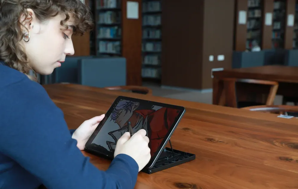 O persoană într-o bibliotecă desenează o ilustrație colorată cu un stilou pe o tabletă așezată pe un suport