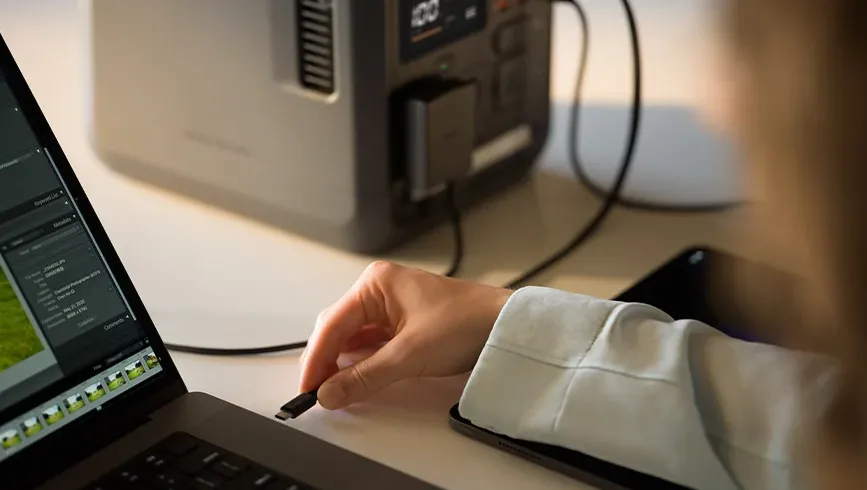 O persoană ține un cablu USB-C negru lângă un laptop cu un program de editare foto deschis, iar în fundal se vede stația de alimentare DJI Power 1000 Mini, conectată la o sursă de alimentare