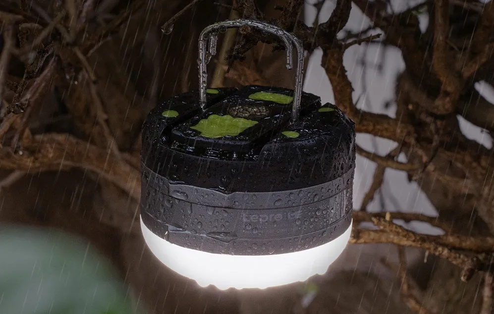 O lampă de camping atârnată de un cârlig metalic printre ramurile copacilor, în timpul unei ploi puternice de seară.