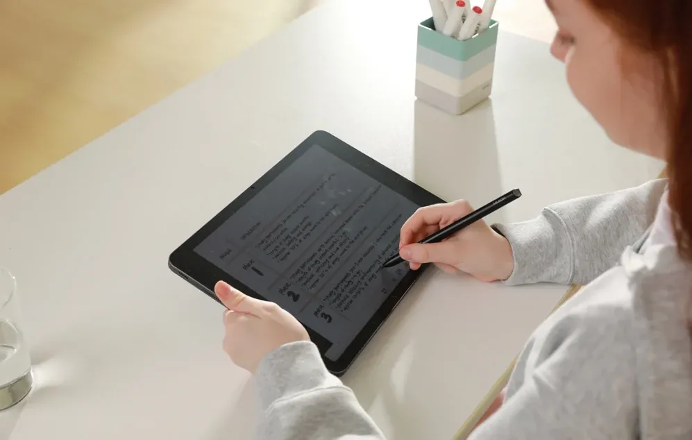 O persoană așezată la o masă albă folosește un stylus pentru a lucra la un document afișat pe o tabletă