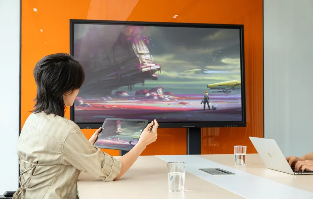 O persoană așezată la o masă ține o tabletă cu un stilou, iar imaginea de pe ecranul acesteia este afișată pe un monitor mare