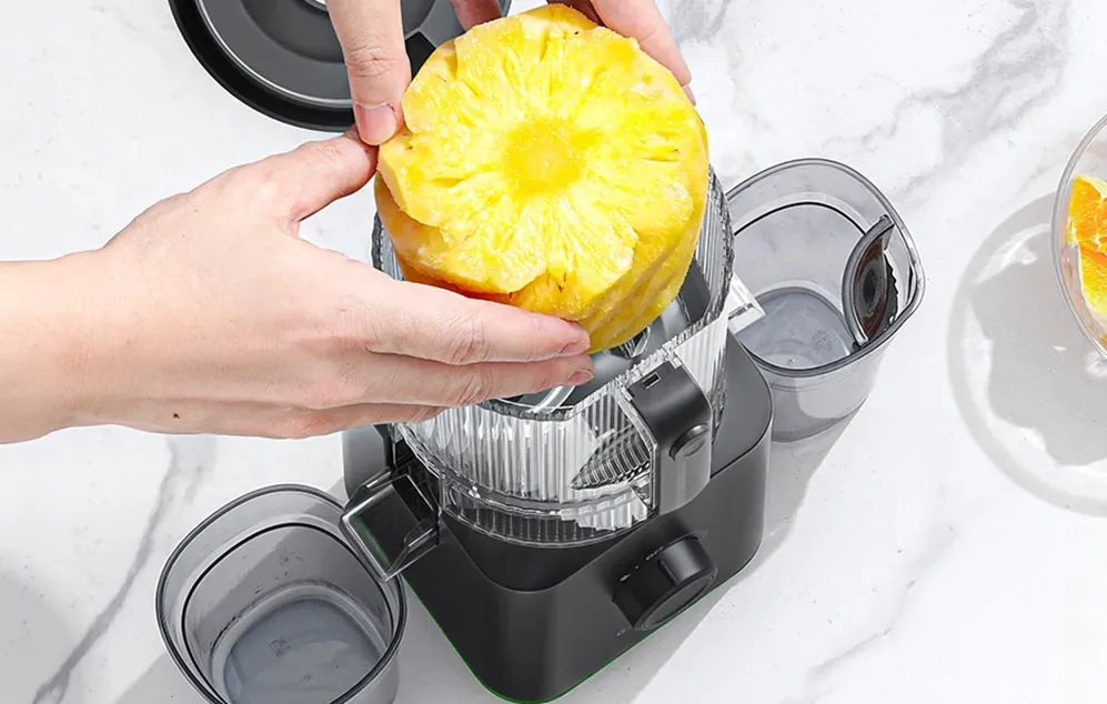 Mâini care introduc o bucată mare de ananas decojit în gura de alimentare a storcătorului lent