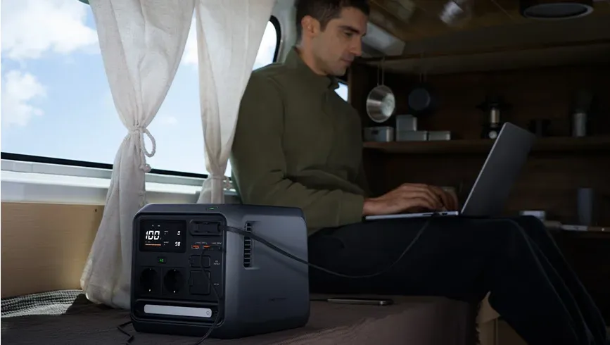 Un bărbat stă în interiorul unei rulote și lucrează la un laptop, care este conectat printr-un cablu la stația de alimentare DJI Power 1000 Mini așezată lângă el pe pat
