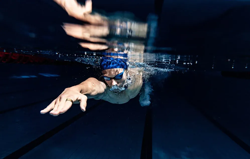 Un bărbat înotând sub apă într-o piscină, cu un inel inteligent auriu purtat pe deget